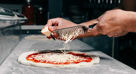 Hands grating cheese onto pizza dough.